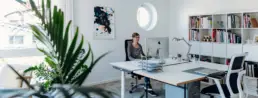 Büro von Studio Somo mit zwei Schreibtischen, Regal und links eine Pflanze, Britta Raab arbeitet am hinteren Schreibtisch