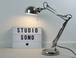 Leuchtschild mit dem Schriftzug Studio Somo und einem Herz wird von einer silbernen Schreibtischlampe angestrahlt