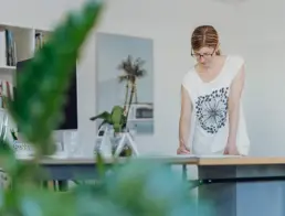 Inhaberin Britta Raab steht am Schreibtisch mit Blick nach unten und einem Stift in der Hand, links im Bild ist eine Pflanze