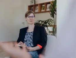 Britta Raab sitzt auf einem Sessel und blickt zu Kundin, von der rechts unscharf die Schulter und die Hand zu sehen sind