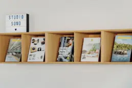 Holz-Regal mit fünf Fächern nebeneinander, darin verschiedene Broschüren, darauf ein Leuchtschild mit dem Schriftzug Studio Somo