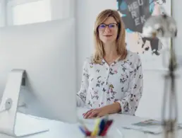 Inhaberin Britta Raab steht hinter ihrem Schreibtisch mit großem Bildschirm und blickt lächelnd in die Kamera,