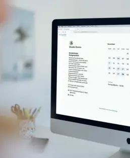 Computer-Bildschirm mit Ansicht eines Terminbuchungs-Tools, links daneben ein Stiftehalter und ein Handy