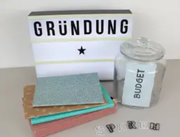 Leuchtschild mit Schriftzug Gründung, Glasgefäß mit Schriftzug Budget, Notizbücher und Buchstaben-Büroklammern mit Schriftzug Sparen