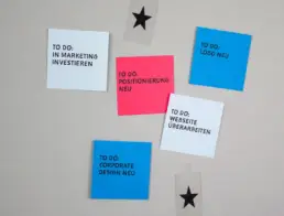 Graupappe-Board mit fünf farbigen Post-its mit Marketing-Aufgaben und zwei schwarzen Sternen