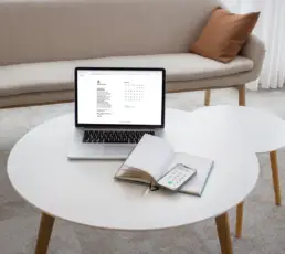 Eine beige Couch und davor zwei runde weiße Tische, darauf ein Laptop zur Terminvereinbarung, ein Kalenderbuch und ein Handy