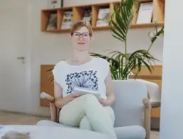 Inhaberin Britta Raab sitzt auf einem Sessel mit einer Broschüre in den Händen und lächelt in die Kamera