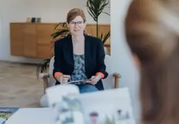 Britta Raab sitzt mit Stift und Notizbuch im Sessel und blickt lächelnd zu Kundin, die unscharf von hinten zu sehen ist